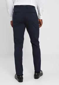 Marineblaue Hose mit geradem Schnitt, zwei Gesäßtaschen mit Knopfverschluss und glatter Oberfläche, kombiniert mit schwarzen Schuhen.