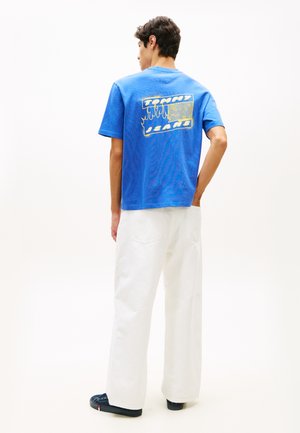 Άνδρας που φοράει φωτεινή μπλε μπλούζα με το γράμμα "Tommy Jeans", λευκό παντελόνι φαρδύ και σκουρόχρωμα αθλητικά παπούτσια, στέκεται μπροστά σε απλό φόντο.