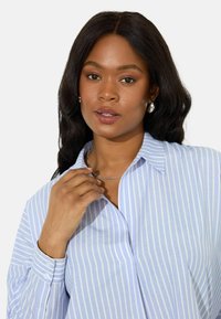 Camicia a righe azzurro chiaro con colletto e maniche lunghe. Presenta una tasca e una morbida texture del tessuto. Collana in catena d'argento.