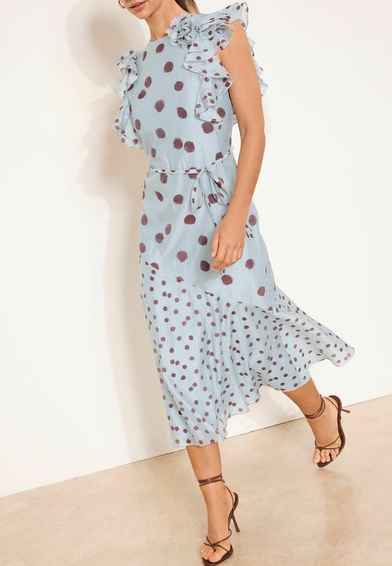 Sieviete valkā gaiši zilu, bezpiedurkņu kleitu ar brūniem punktiņiem, volānu uz pleciem un asimetrisku apakšmalu, kopā ar brūnām sandales ar siksniņām.