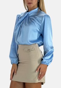 Hellblaue Satinbluse mit puffigen Ärmeln und einem gebundenen Kragendetail. Kombiniert mit einem beigen Minirock mit zwei Fronttaschen und einem Knopf.