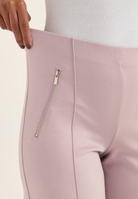 Nahaufnahme einer Person, die den Taillenbund einer hellrosa Hose mit einem vertikalen silbernen Reißverschluss an der Seite dehnt.