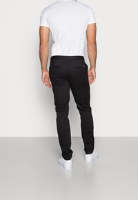 Pantaloni neri slim-fit con una texture liscia e quattro tasche, indossati con una t-shirt bianca aderente e sneakers bianche.