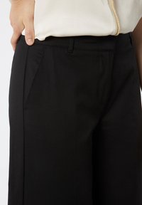 Pantalones de pierna ancha negros con una textura suave, que cuentan con bolsillos delanteros y una cinturilla ajustada. Parte de una blusa de color claro es visible por encima.
