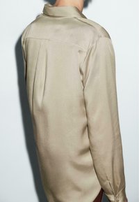 Camicia beige a maniche lunghe realizzata in tessuto liscio, con un colletto classico, pieghe posteriori e polsini abbottonati, dal taglio morbido.