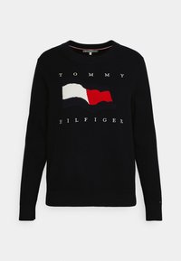 Maglione blu navy con colletto rotondo, caratterizzato da un design di bandiera ricamata in rilievo nei colori rosso, bianco e blu. Testo del logo sottostante in bianco.