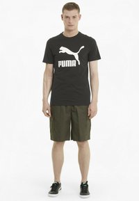 Must värvi T-särk valge Puma logo ja tekstiga, millega on sobitatud oliivroheline cargo lühikeste pükste paari. Mudel kannab musti tossusid valgete talladega.