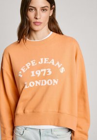 Oranžs džemperis ar baltu tekstu "PEPE JEANS 1973 LONDON." Apaļš kakls, garas piedurknes un brīvs piegriezums. Tekstūras auduma detaļa.