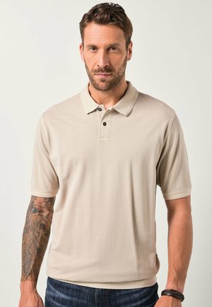 Hombre con cabello corto castaño y barba, vistiendo una camiseta polo beige y jeans, mostrando el antebrazo izquierdo tatuado sobre un fondo claro.