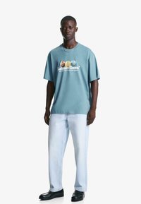 Svetlo modra oversize majica s tekstom "Leisure Game" in pisanymi grafičnimi žogami. Ujemite jo z svetlim denim hlačami in črnimi čevlji.