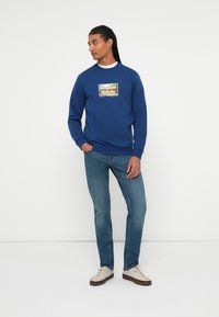 Sudadera azul con un estampado gráfico en la parte frontal, combinada con jeans de un azul claro y zapatillas de color crema. Tejido suave y ajuste casual.