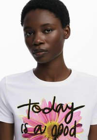 Witte katoenen t-shirt met een grote roze madelief grafiek met gele accenten en geborduurde tekst die leest "vandaag is een goede."