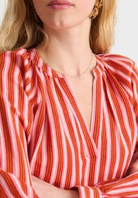 Gestreifte pinke Bluse mit vertikalen roten, orangen und hellpinken Linien, V-Ausschnitt, gerafften Schulternähten und langen Ärmeln.