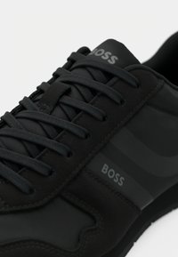 Ténis preto com material texturizado, apresentando atacadores escuros, logótipo na lateral e riscas subtis como detalhe de design. Sola em borracha.