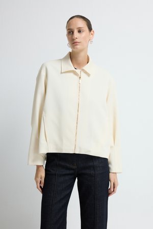 Crème geknipte jas met een kraag, een ritssluiting aan de voorkant en lange mouwen, gedragen met donkerdenim broek, gladde textuur.