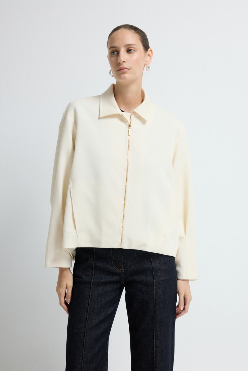 Crème geknipte jas met een kraag, een ritssluiting aan de voorkant en lange mouwen, gedragen met donkerdenim broek, gladde textuur.