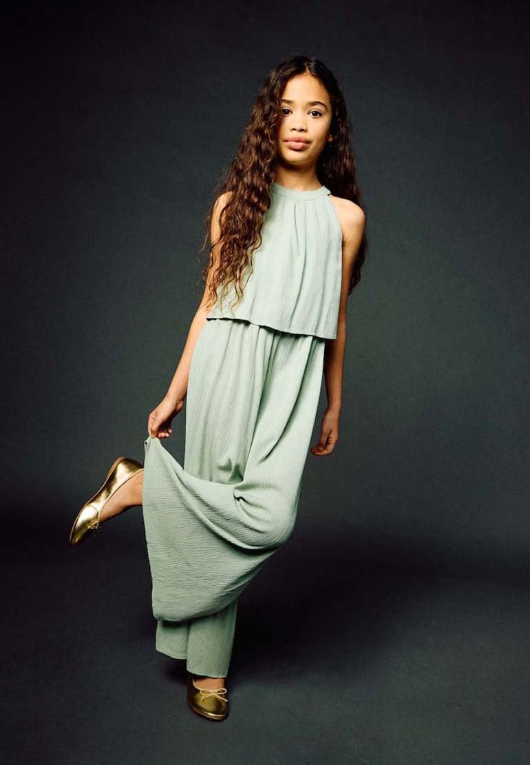 Fille aux longs cheveux bouclés vêtue d'une combinaison sans manches vert pâle soulevant une jambe de pantalon, portant des ballerines dorées sur fond sombre.