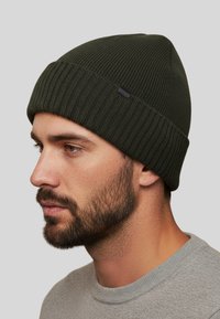 Beanie verde lavorato a maglia con texture a coste, cima arrotondata e una piccola etichetta nera. Indossato con una felpa grigia a girocollo.