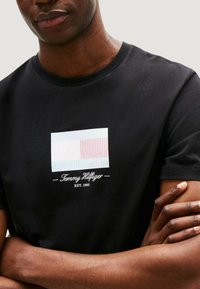 Czarny T-shirt z prostokątnym logo w niebieskie i czerwone paski oraz białym tle; "Tommy Hilfiger EST. 1985" w białym tekście poniżej.