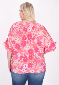 Blusa floreale con base rosa, fiori bianchi e arancioni, maniche corte a volant, vestibilità comoda e dettaglio a chiave sulla parte posteriore.