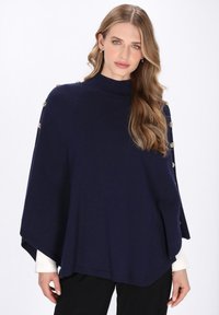 Poncho albastru marin cu gât înalt, decorat cu butoane aurii pe laterale, textură netedă și croială lejeră, purtat peste o cămașă albă.