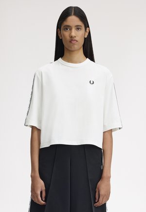 Ung kvinde med langt mørkt hår iført en hvid T-shirt med korte ærmer og logo på brystet samt sort plisseret nederdel mod en ensfarvet baggrund.