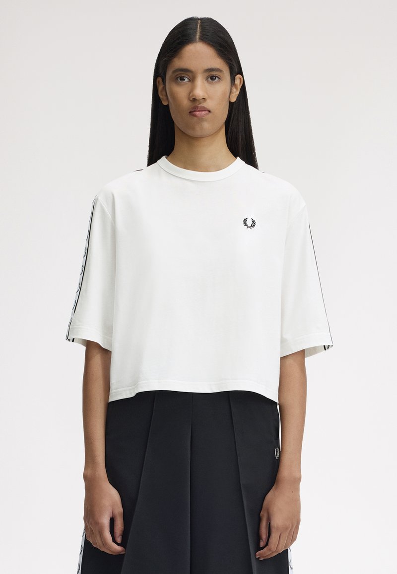 Jeune femme aux longs cheveux bruns portant un t-shirt blanc à manches courtes avec un logo sur la poitrine et une jupe plissée noire, sur un fond uni.