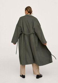 Lang, oversized grijs jasje van een wolmix, met een ceintuur in de taille, brede mouwen en zijzakken, gestyled met beige broeken en zwarte schoenen.