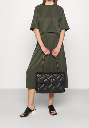 Borsa trapuntata nera con tracolla a catena dorata e dettaglio della chiusura, accanto a una modella con un vestito largo verde oliva e sandali neri con tacco.