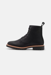 Bottes de cheville en cuir noir avec une finition texturée, un design à lacets, une semelle en caoutchouc et un bord contrasté tan le long de la semelle.