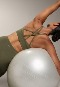 Športni modrček olivno zelene barve s prečnimi naramnicami, kombiniran z visokimi legicami v istem odtenku. V ospredju je odsevna telovadna žoga.