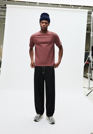 Piros bordázott rövid ujjú ing, laza fekete farmer, szürke tornacipő. A modell sötétkék kötött sapkát visel. Egyszerű stúdió háttér.