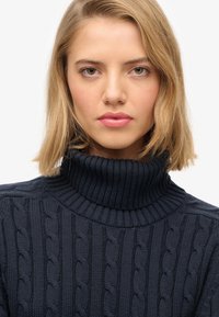 Mujer rubia con cabello liso, vistiendo un suéter de cuello alto de punto trenzado de color azul oscuro, mirando directamente a la cámara con una expresión neutral.