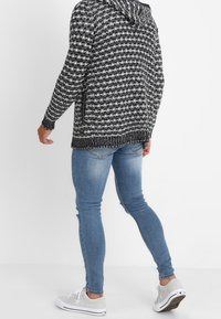 Schwarz-weißer, gestrickter Hoodie mit Kapuze und strukturiertem Muster, kombiniert mit hellblauen Jeans und grauen Turnschuhen.