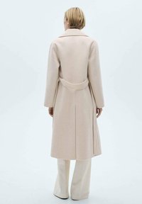 Abrigo color beige claro con un diseño estructurado, con un cuello ancho, cinturón en la cintura y un detalle de costura central en la parte posterior. Textura de tela suave.