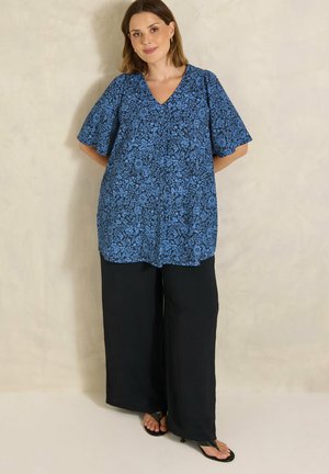 Vrouw draagt een blauwe bloemenblouse met V-hals en wijde mouwen, losse zwarte wijd uitlopende broek en zwarte sandalen, staand tegen een effen muur.
