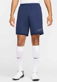 Dunkelblaue Sportshorts mit elastischem Bund, versehen mit einem weißen Nike-Logo auf dem linken Bein. Kombiniert mit weißen Kniestrümpfen und Fußballschuhen.