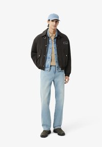 Chaqueta bomber negra sobre una chaqueta de mezclilla, camisa beige, jeans azul claro y zapatillas grises. También lleva una gorra azul claro.