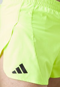 Neon grønne sports shorts med elastisk talje, der har sideslids og et sort tre-stribe logo ved kanten.