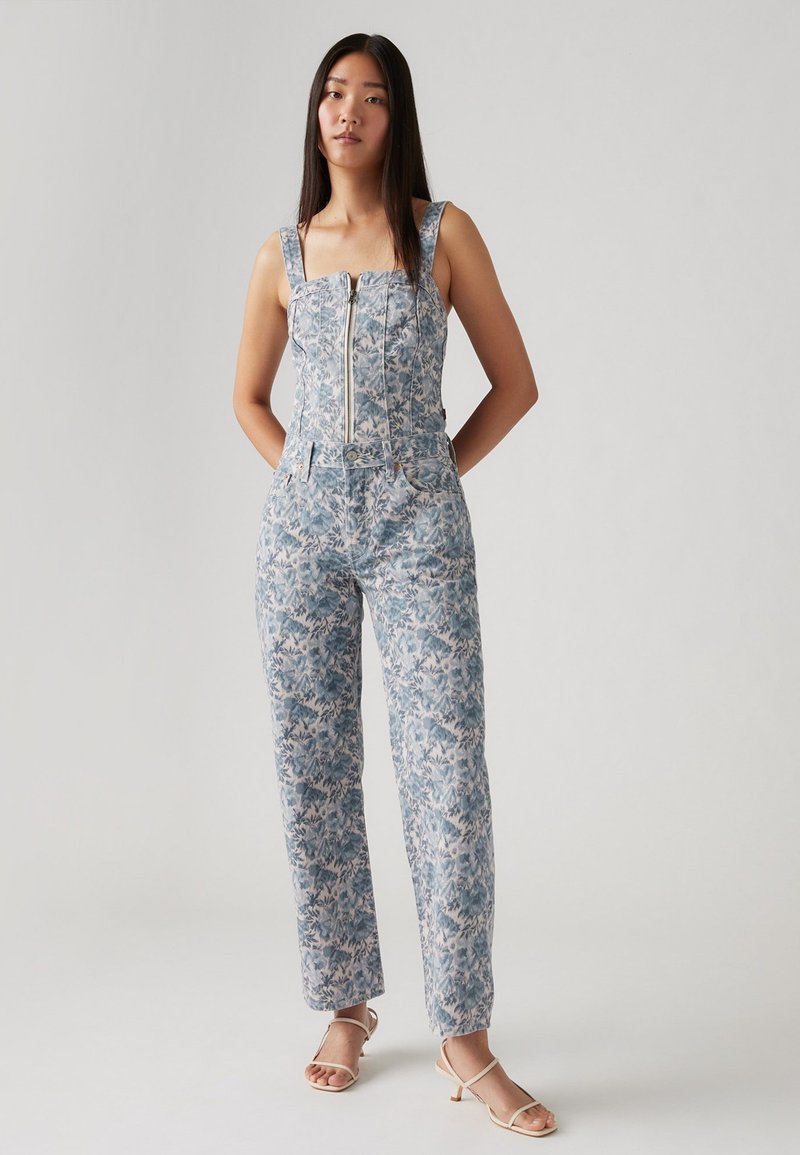 Combinaison florale présentant des motifs bleus et blancs, un corsage structuré avec une fermeture éclair à l'avant, de larges bretelles et une silhouette à jambes droites.