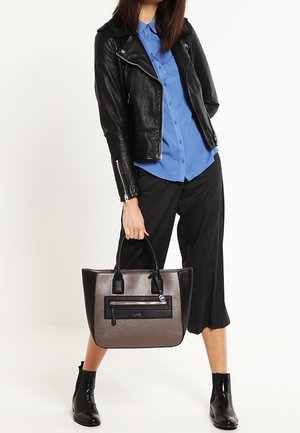 Veste de moto en cuir noir portée sur une chemise bleue à boutons. Modèle portant un sac à main structuré gris avec des accents noirs et une poche zippée, chaussé de bottines noires.