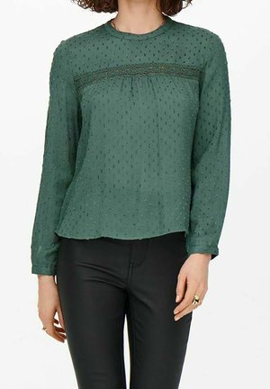Blusa verde a maniche lunghe in tessuto testurizzato con un pannello in pizzo decorativo sul busto. Accentuata da piccoli punti in rilievo.