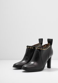 Botines de cuero negro hasta el tobillo con punta afilada, ojetes dorados y un tacón texturizado alto. Presentan un cordón decorativo a lo largo del borde superior.
