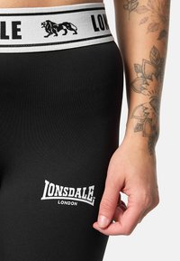 Black leggings with a white logo featuring "LONSDALE LONDON." The waistband is white with a black lion design and text. Smooth texture.