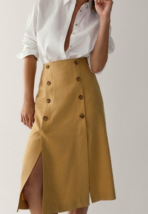Femme portant une jupe taille haute jaune moutarde avec des boutons et une fente sur le devant, associée à une chemise blanche boutonnée avec des manches retroussées.