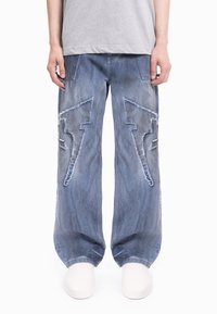 Modre široke kavbojke iz jeansa z raztrganimi detajli in velikimi vzorci strele na vsaki nogi, z zbledele zaključkom in sproščenim krojem.