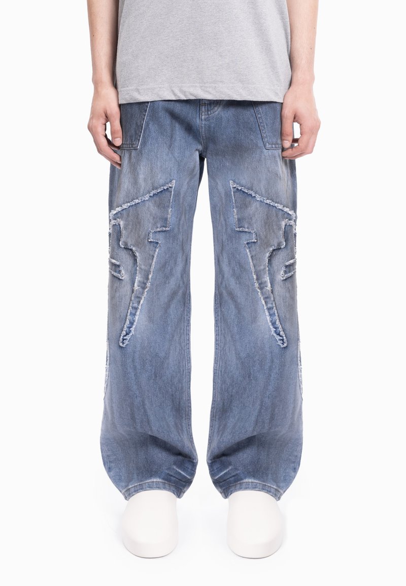 Modre široke kavbojke iz jeansa z raztrganimi detajli in velikimi vzorci strele na vsaki nogi, z zbledele zaključkom in sproščenim krojem.
