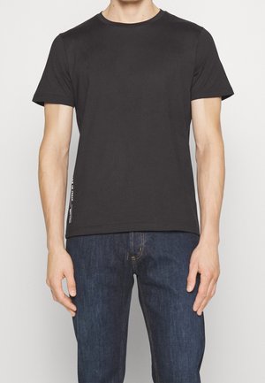 Homme portant un t-shirt noir uni à manches courtes et un jean bleu foncé, debout devant un fond clair.