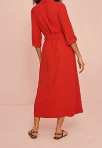 Femme portant une longue robe rouge ceinturée avec des manches retroussées et des sandales marron, se tenant devant un fond beige uni.