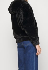 Persona che indossa una giacca a cappuccio in peluche nera e jeans di denim blu, vista da dietro su uno sfondo neutro.